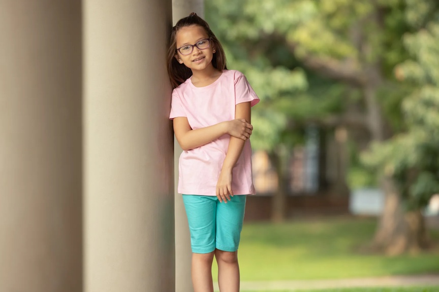 A young girl wearing glasses leaning against a column, smiling in a park setting with trees in the background.