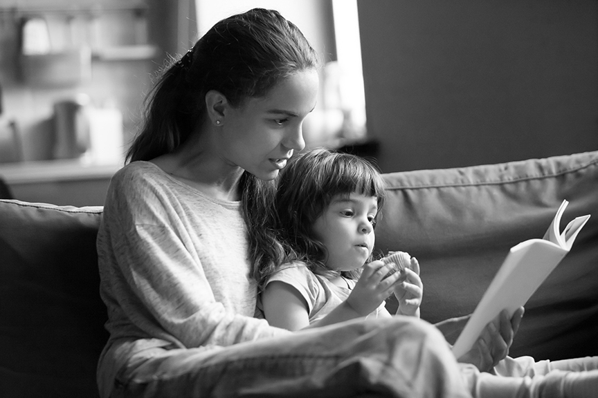 A mom sits on a couch at home, reading a book with her young preschool aged daughter seated next to her. The daughter is holding a snack.