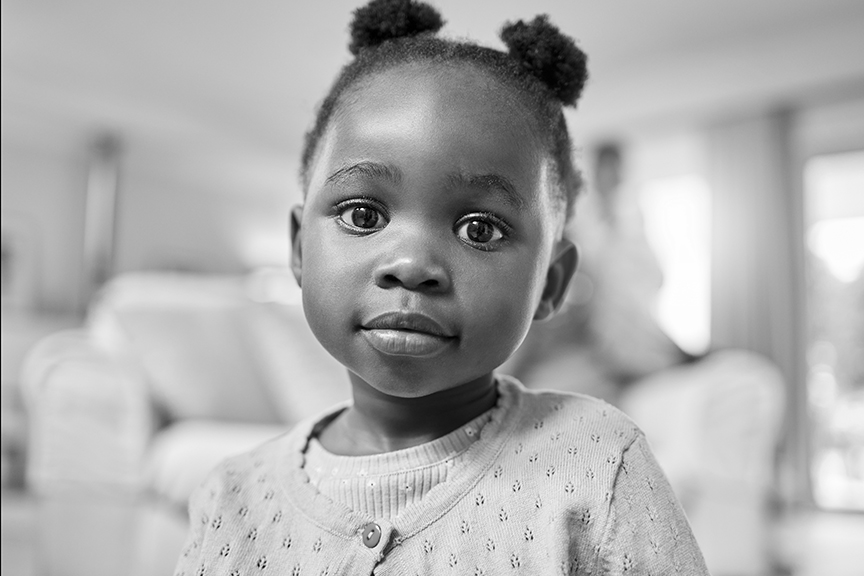 A preschool aged girl is wearing a cardigan and is shown in her home. She's looking at the camera directly.