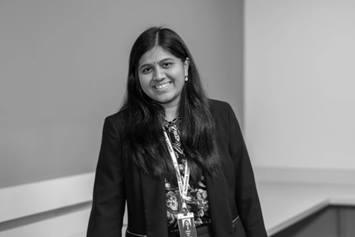 Dr. Aluri is wearing a dark blazer. She's smiling, standing in an office setting with a desk behind her.