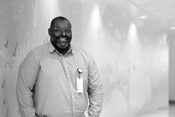 Jay Hudgins is smiling, wearing a long-sleeve button down shirt. He's standing in a hallway, with tree and bird graphics on the wall behind him.