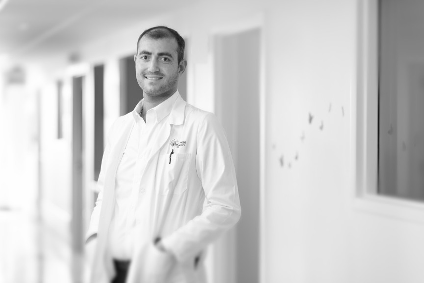 Dr. Qussous is smiling, standing in a hallway, wearing a white coat with a white buttoned shirt underneath. 