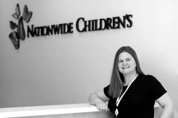 Jessica is standing, wearing a dark short sleeve shirt. Her right elbow is resting on a desk. The Nationwide Children's logo is on the wall behind her.