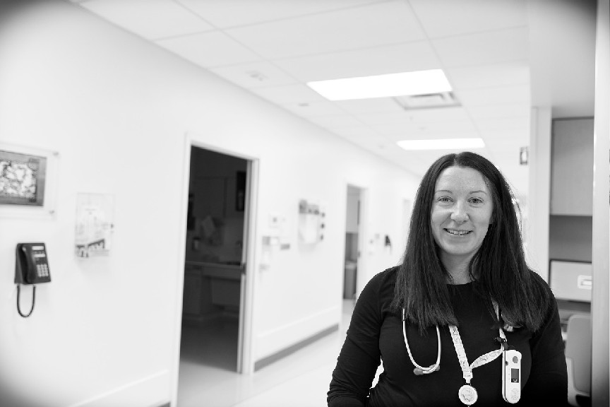 Joanne is smiling, standing in a clinical hallway with patient rooms in the background.