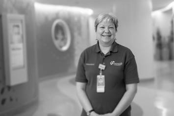 Karen is standing in a hallway, smiling. She's wearing a volunteer polo and a name badge. Tree and forest graphics are on the wall behind her.