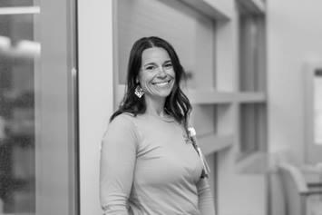 Katie is wearing a solid long sleeve shirt and statement earrings. She's smiling, standing in a waiting room environment.