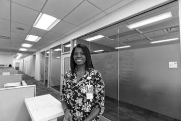 Tifini is standing in an office setting, with frosted glass panes behind her. She's smiling and wearing a patterned shirt.