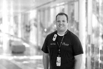Zachary is wearing a dark t-shirt, smiling, standing in a light-filled hallway.