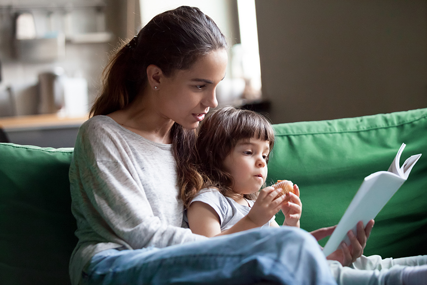 A mom sits with her young daughter, who is not feeling well, on a kelly green couch. Mom is reading to the daughter as the little girl eats a muffin.