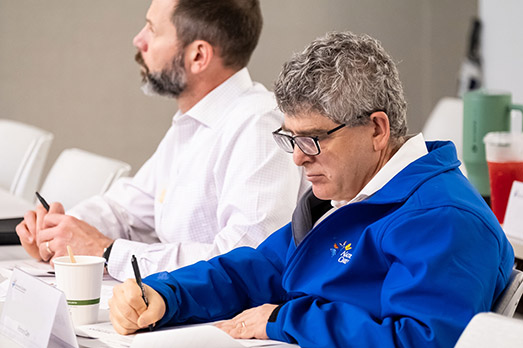 Two professionals seated at a table taking notes during a meeting, with one wearing a blue jacket and focusing on writing.