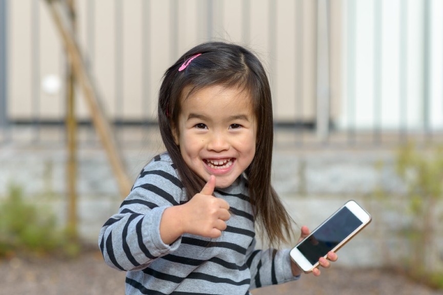 Adorable young Chinese girl smiling for a photo with a cell phone in one hand and a thumbs up in the other.