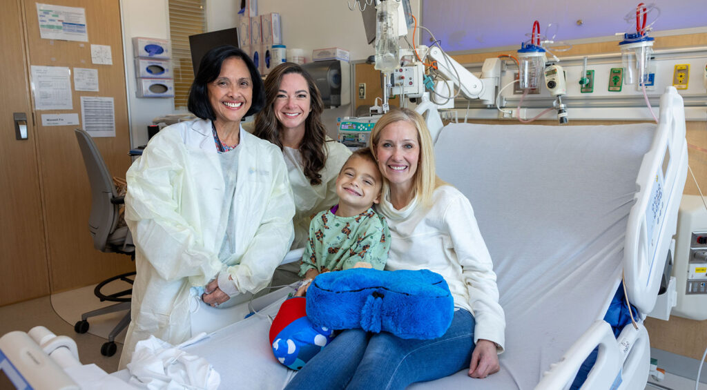 A child sits upright in a hospital bed holding a blue plush pillow, surrounded by three adults wearing protective gowns in a brightly lit hospital room with medical equipment and an IV setup visible behind the bed.