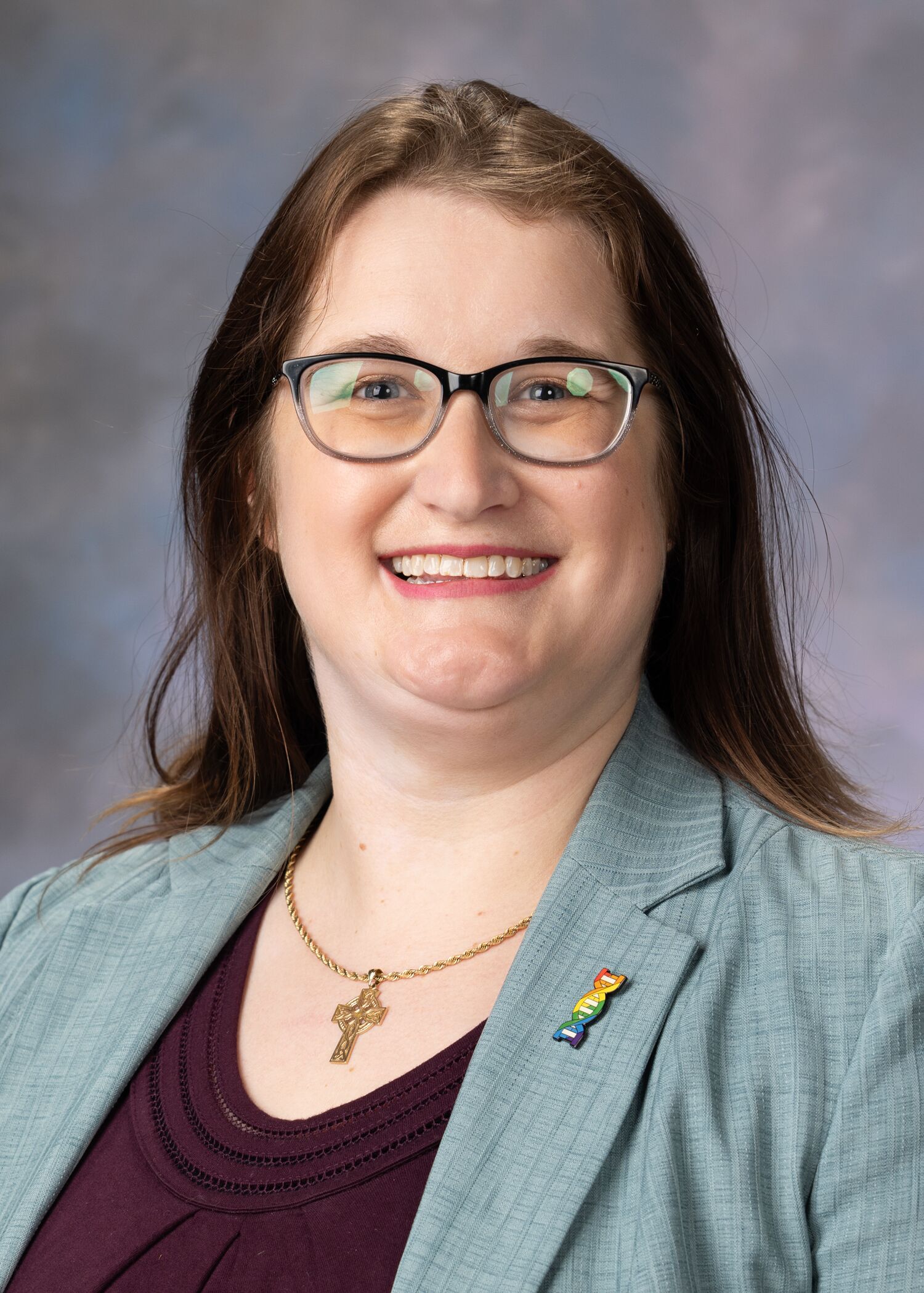 Portrait of Nettie Pyne. Nettie is wearing glasses, a cross necklace, blue jacket and a maroon shirt. 