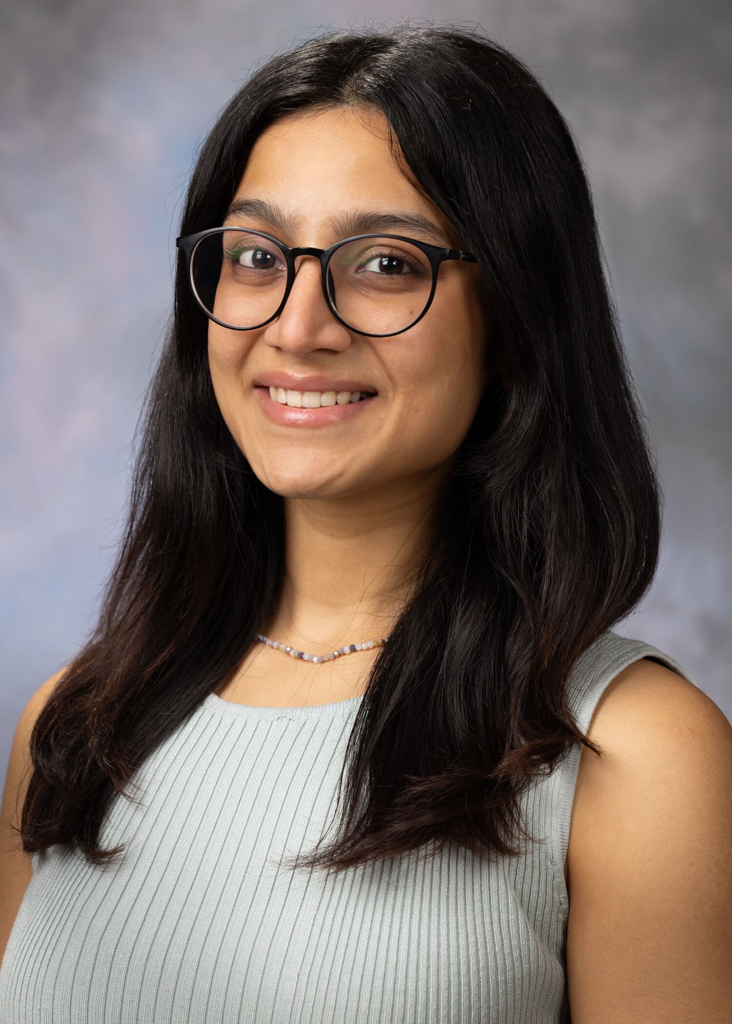 Portrait of Dipti Pant. She has medium length, dark brown hair, a light-colored tank top and dark rimmed glasses. The background is a mix of grays and pinks.