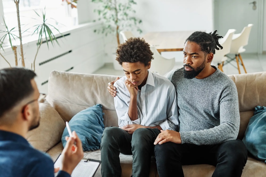 Father with his teenage son at meeting with social worker, psychologist discussing mental health family sitting on sofa in psychotherapist office