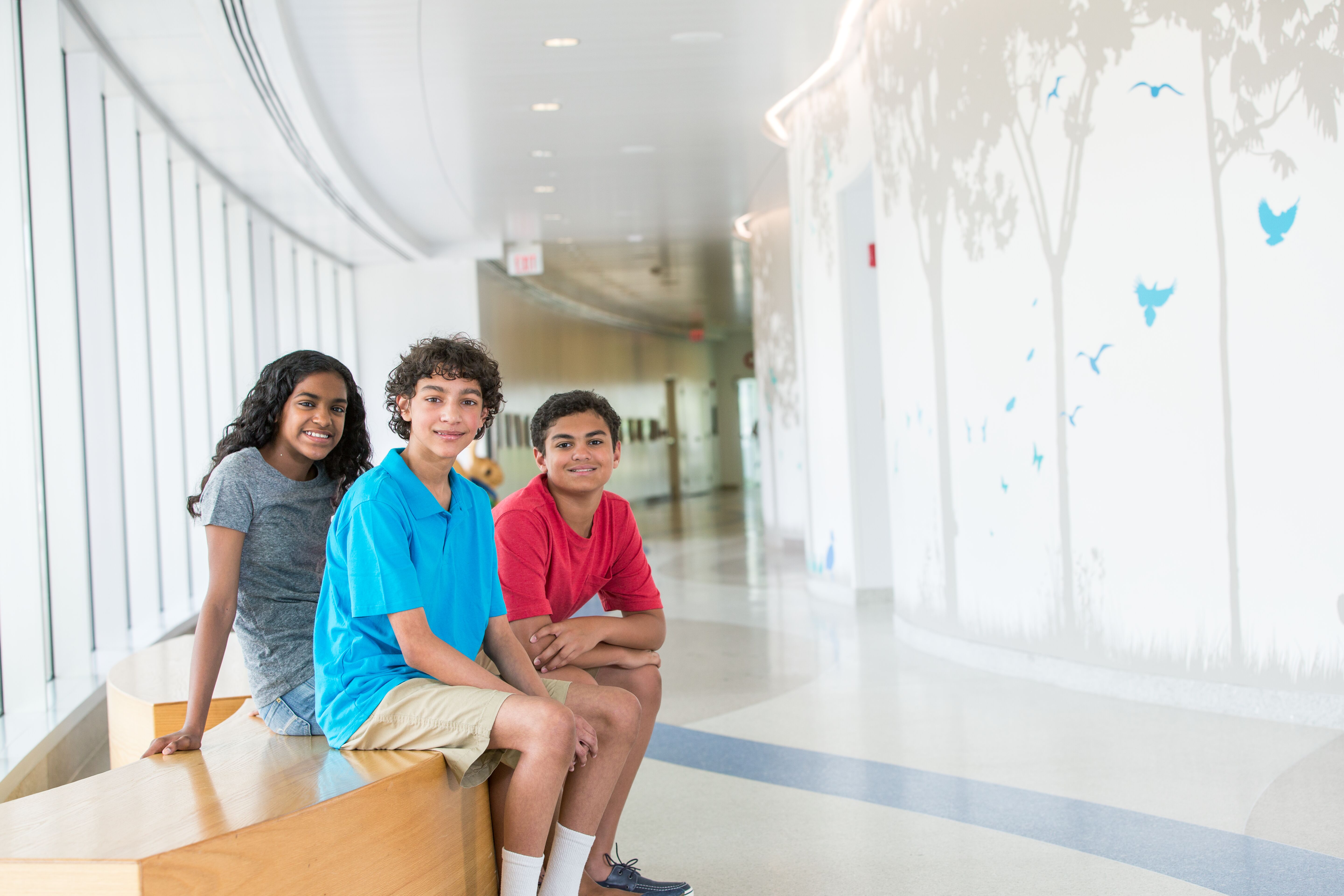 Three children sitting on a bench in a hospital corridor, smiling and engaged in conversation, with colorful wall murals in the background.