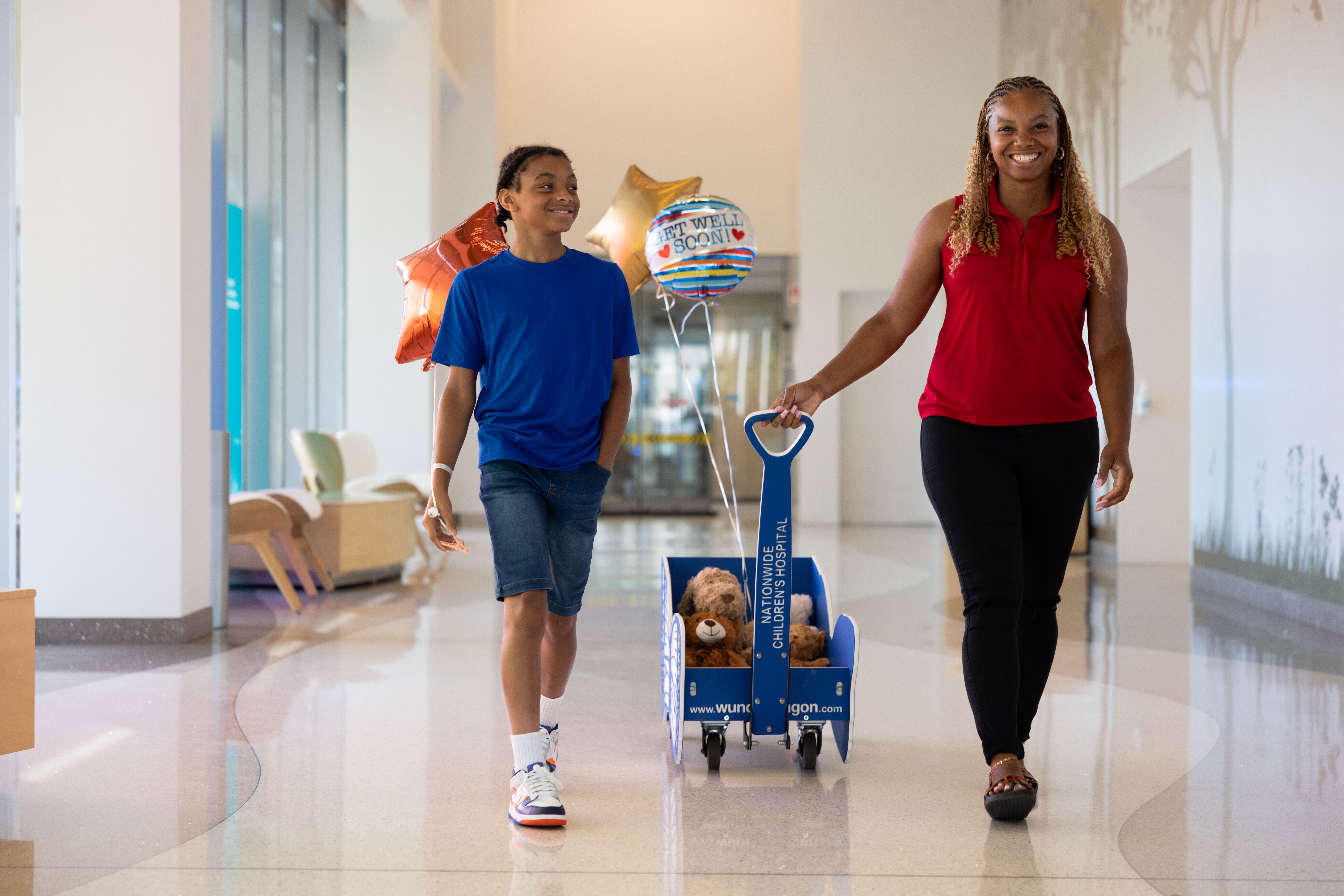 A woman and child walk through a hospital corridor, the child pulls a wagon with a stuffed animal and balloons.