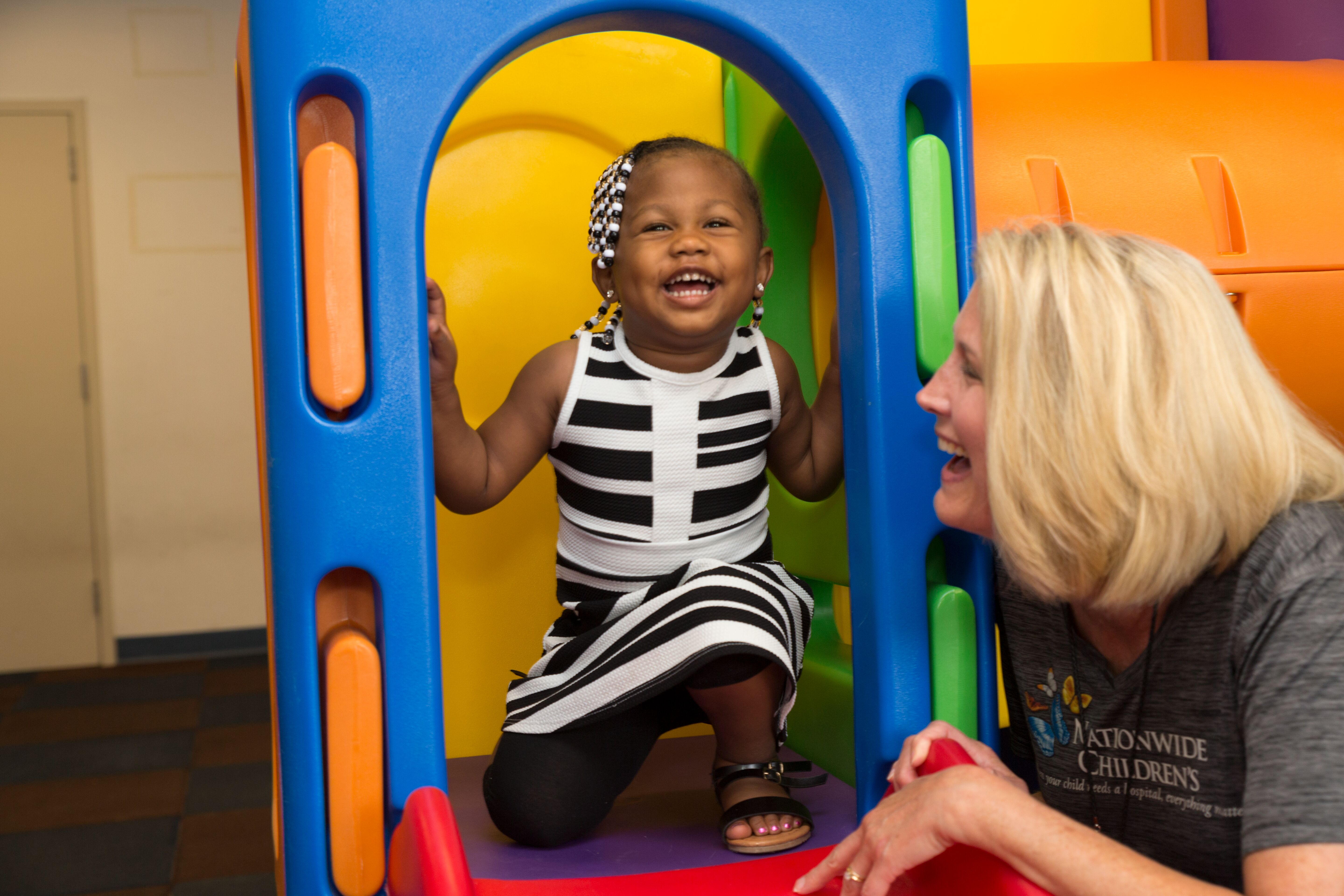 A young girl smiles joyfully while playing on a colorful play structure, with an adult nearby watching her happily.