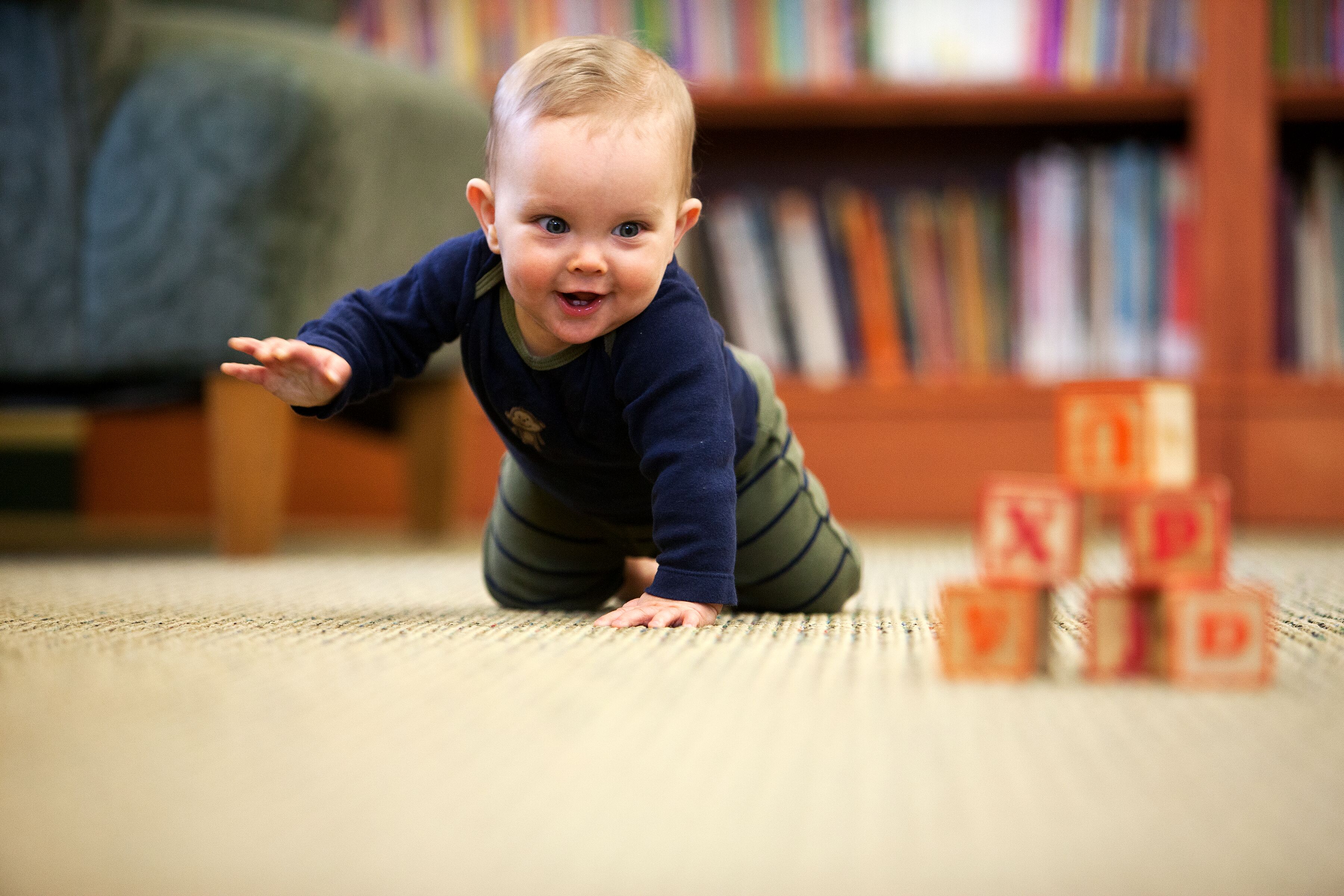 A baby crawling towards a stack of colorful blocks on a carpeted floor in a library setting.
