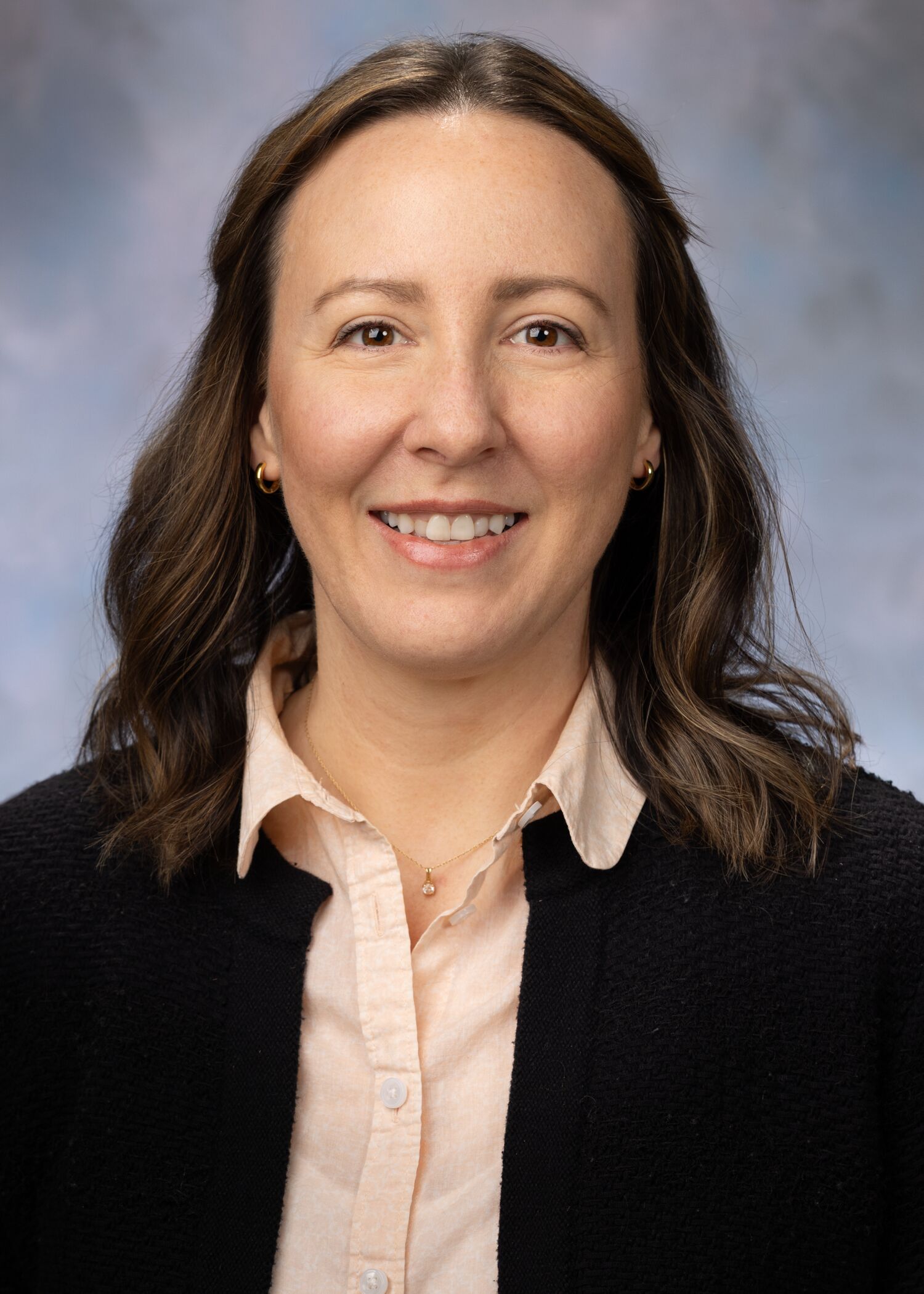 Portrait of Lauren Sebben. Lauren has shoulder-length brown hair and is wearing a pink button up shirt with a black jacket. 