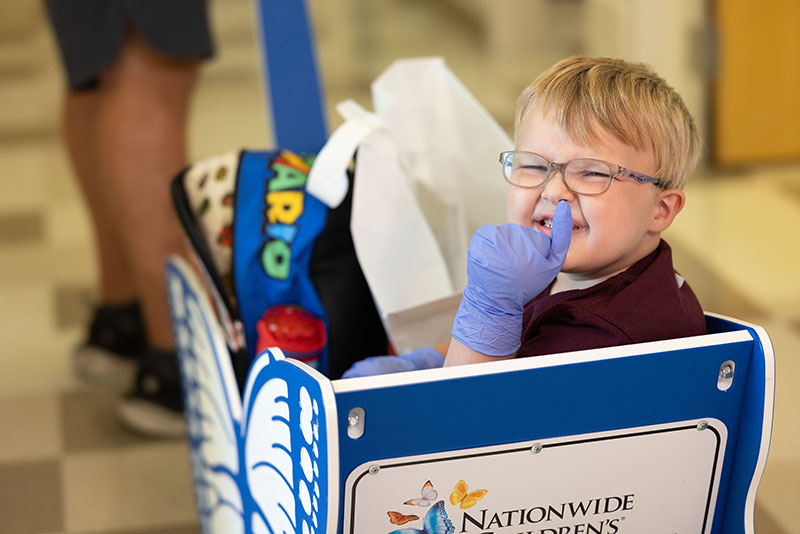A young boy wearing glasses smiles as he rides in a blue and white wagon.