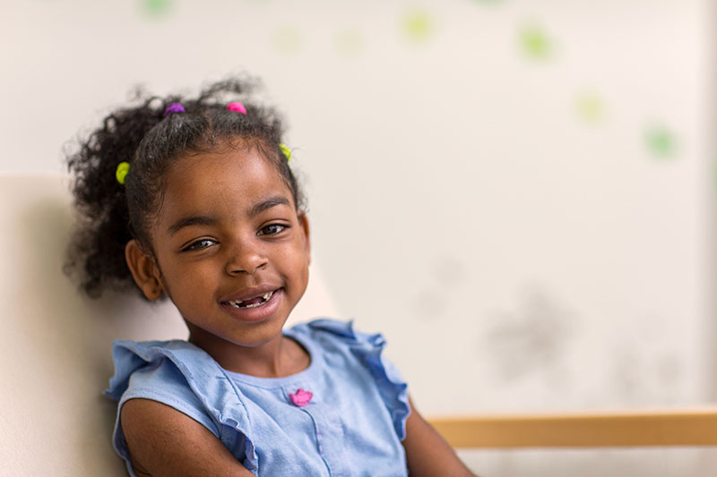 A young girl wearing a blue dress smiles as she looks at the camera.