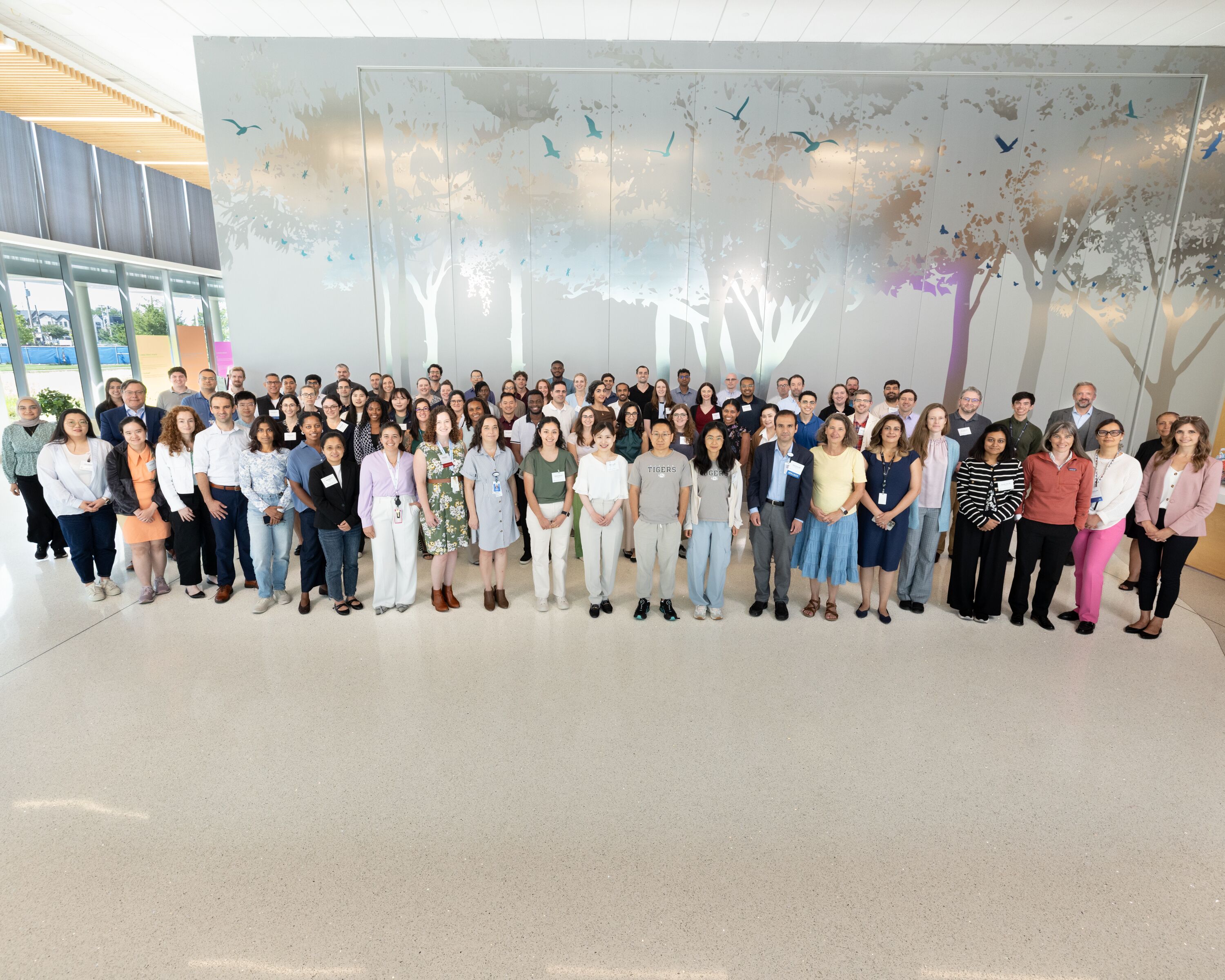 Large group photo of approximately 80 to 100 people standing together in a bright, modern lobby. Behind the group is a floor-to-ceiling wall mural featuring stylized trees and flying birds in soft gray, white, and blue tones. The space has high ceilings, large windows along the left side, and a polished light-colored floor. Most individuals are wearing business casual attire and name badges, arranged in several rows facing the camera. Natural light fills the open interior, creating a professional and welcoming setting.