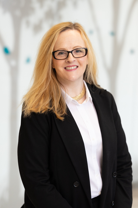 Photo of Susannah Wolman. Susannah has shoulder-length light-colored hair wearing a black blazer over a white collared shirt and a gold necklace, standing indoors against a softly blurred background with tree-like shapes.