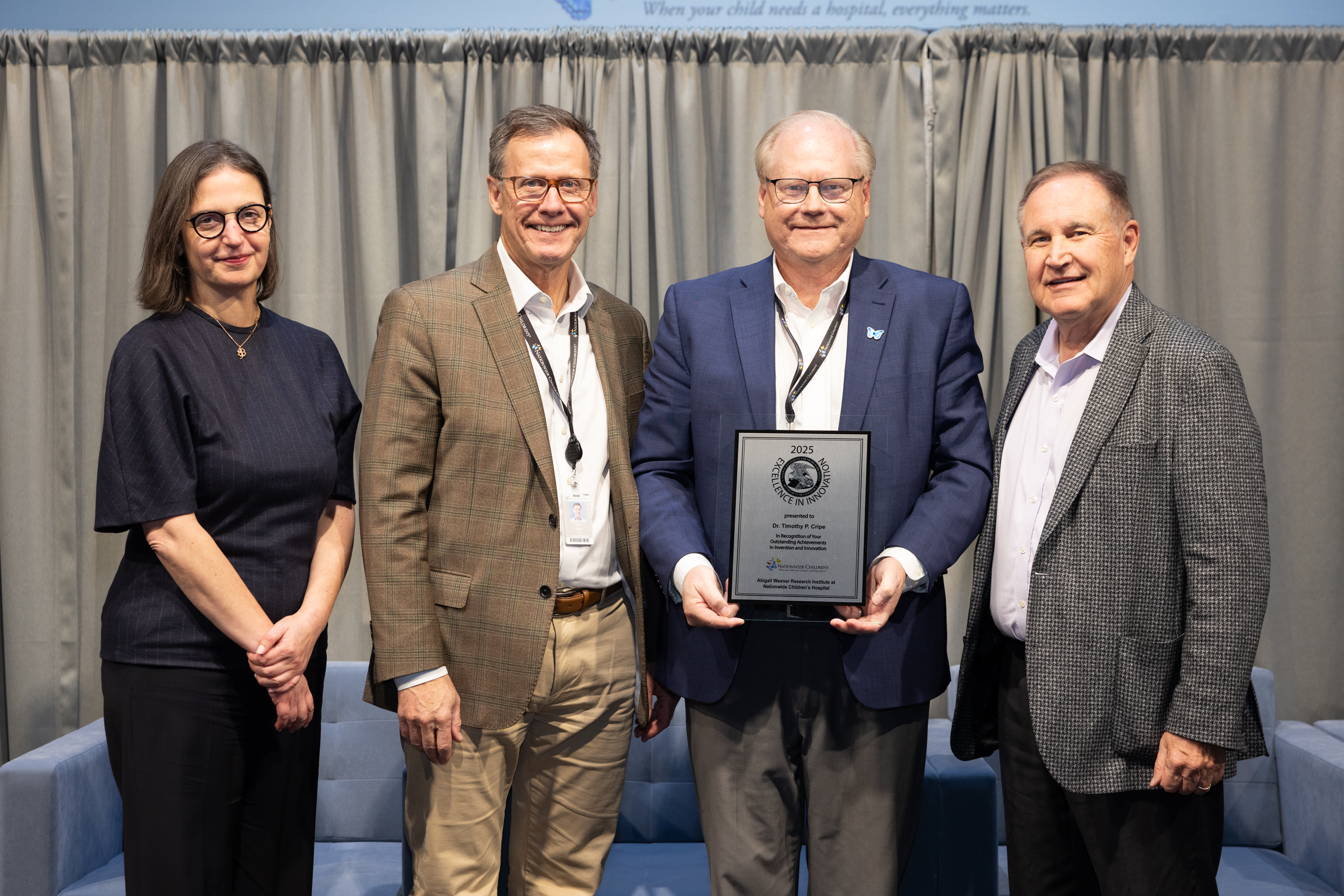 Four individuals stand together indoors in front of a curtain backdrop. The individual in the center is holding a large rectangular award plaque with both hands. The group is dressed in business or professional attire, and they are positioned closely together for a posed photo. Two upholstered chairs are visible at the edges of the scene.