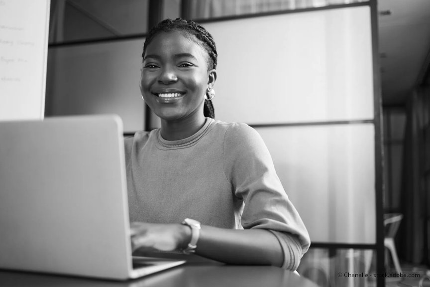 A professional woman is smiling, seated at her laptop in an office setting.