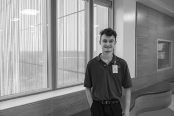 Project SEARCH employee Layne is shown wearing a polo shirt and badge. Layne is smiling and standing in front of a wall of windows.