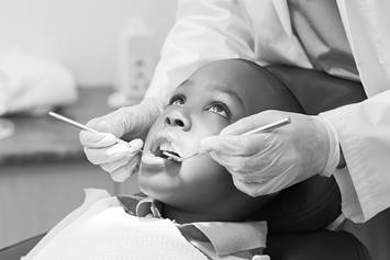An elementary aged child is sitting in a dental chair. His mouth is open and a dental hygienist is holding dental tools near his mouth.