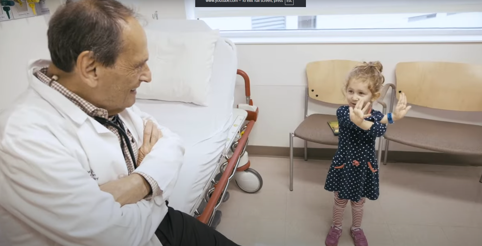 Photo of doctor sitting on bed looking at small child standing on the floor. 
