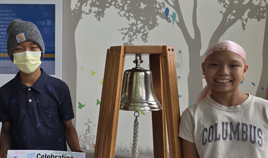  Two individuals standing indoors beside a large silver bell mounted on a wooden frame. The bell has a thick braided rope hanging from its clapper. The person on the left is wearing a dark short-sleeve shirt with a subtle pattern and a knitted gray hat. The person on the right is wearing a light-colored T-shirt with the word “COLUMBUS” printed in bold letters and a light pink head covering. In the background, there is a wall mural featuring silhouettes of trees and colorful butterflies, along with a blue and white poster partially visible on the left side. The setting appears bright and welcoming.