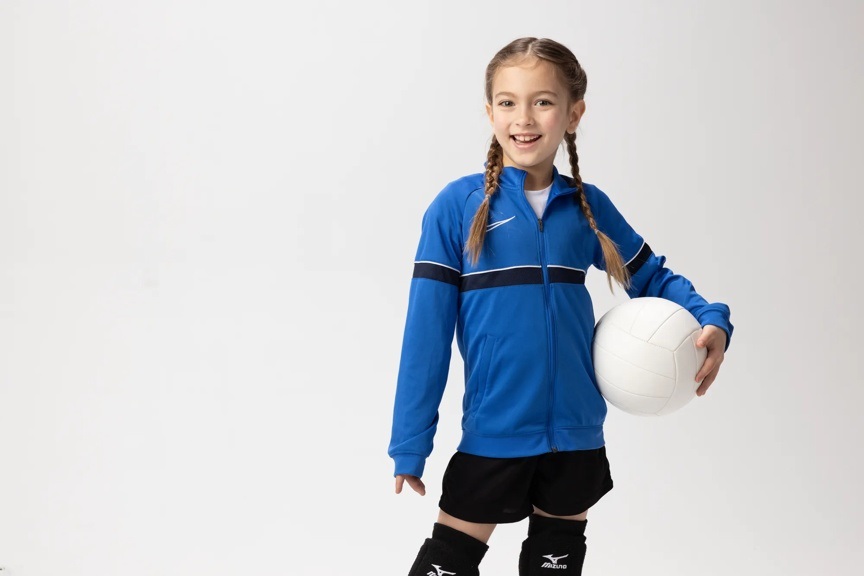 Young girl wearing braids and a blue sweatshirt is smiling at the camera and holding a volleyball against her hip.