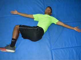 Athletic trainer lays on the ground with knees together and turned to the side. Arms stretched out on each side.