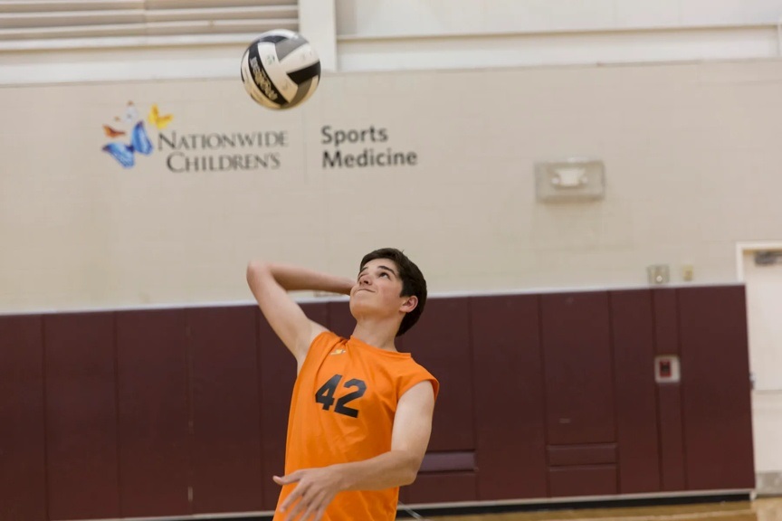A young high school boy is serving a volleyball that has been thrown in the air. He is wearing an orange jersey with the number "42" on it in black numbers.