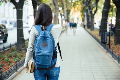 A teenage girl has her back to the camera. She's wearing a back pack and is walking on a path in a tree-lined public area.