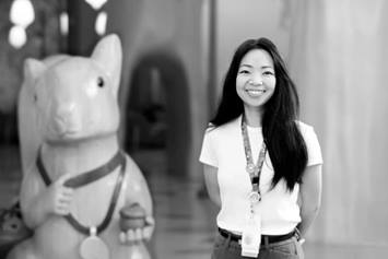 Rebecca has long, dark hair and is wearing a light tee shirt. She's standing in a hallway with a large carved wooden squirrel in the background behind her.